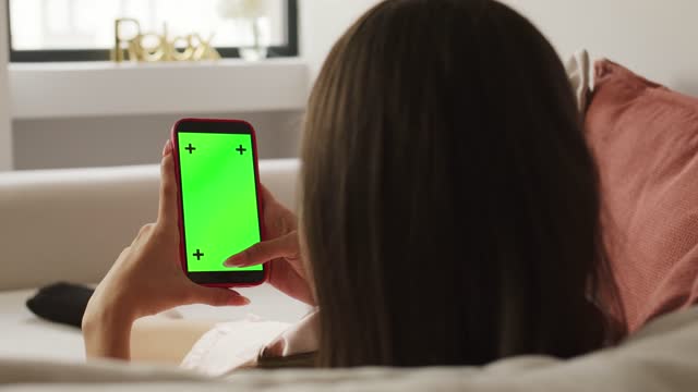 A young woman relaxing on the couch with mobile phone device featuring a green screen .