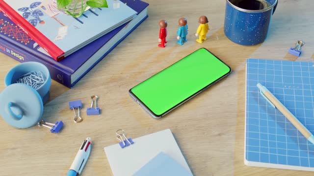 Mobile phone featuring a green screen surrounded by several books, paper clips, binder clips, a blue notebook with a grid pattern, a pen, and a cup.