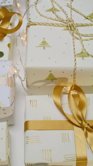 Vertical video of an assortment gifts wrapped with elegant with Christmas paper and golden ribbons over a white surface.